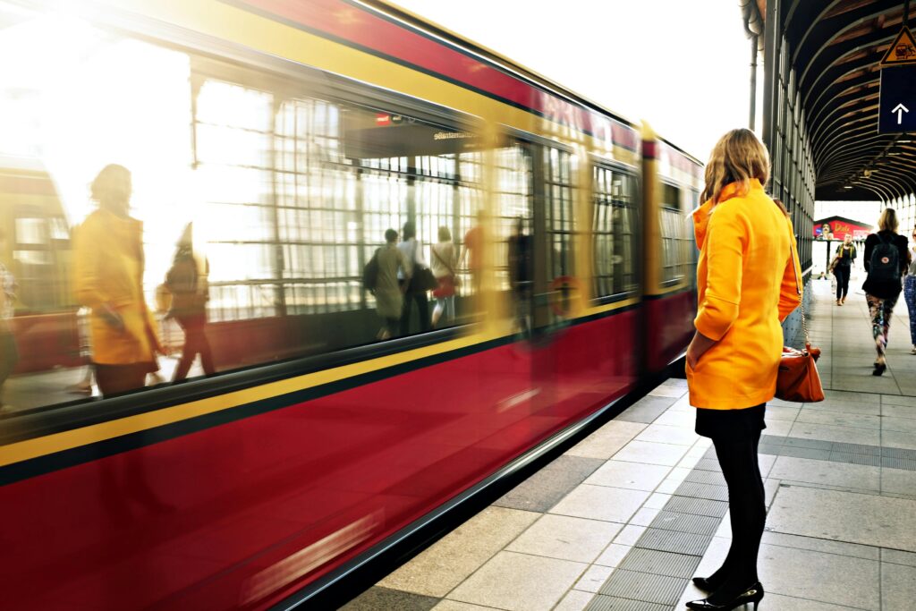 A woman in a yellow coat waits as a red train passes in a bustling station.